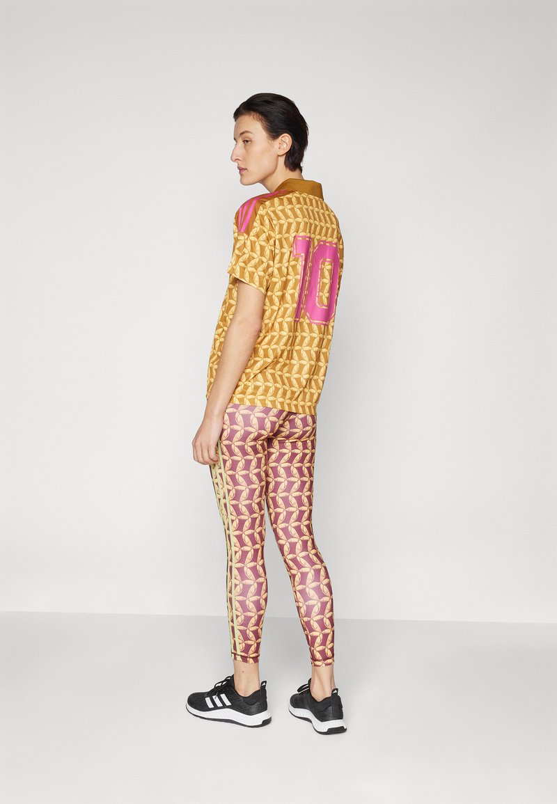 Camicia gialla a fantasia con accenti rosa e numero 10, abbinata a leggings viola con un motivo simile e strisce gialle laterali. Sneakers nere.