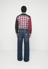 Camicia a quadri con motivi e colori vari (rosso, verde) abbinata a jeans a gamba larga blu con un design a stella sulla tasca posteriore.