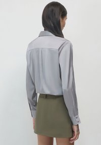 Camicia lunga grigia lucida con colletto a punta, piega sul retro e polsini abbottonati, abbinata a una gonna aderente verde oliva.