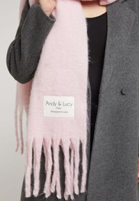 Écharpe en laine rose clair avec des bords frangés, ornée d'une étiquette blanche sur laquelle est brodé "Andy & Lucy Paris Wrapped in Love". Portée avec un manteau gris.