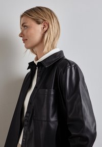 Zwarte leren shirtjacket met een kraag, voorzak en knoopsluiting, gelaagd over een witte turtleneck. Glad oppervlak.