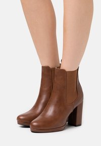 Bottines en cuir marron avec des panneaux élastiques sur les côtés, bout arrondi et un talon bloc. Texture lisse et détails cousus.