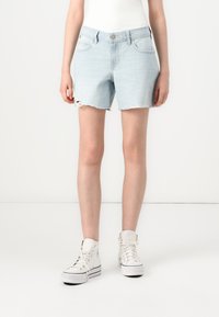 Bermuda de ganga azul clara com uma bainha desfiada, apresentando passantes para cinto e bolsos frontais, combinada com ténis brancos de cano alto.