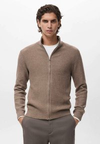 Giovane uomo con capelli ondulati che indossa un maglione zip-up a coste beige sopra una camicia bianca e pantaloni grigi, mani nelle tasche, espressione neutra.