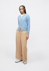 Ανοιχτό γαλάζιο πλεκτό ζακέτα με V-λαιμόκοψη και κουμπιά, συνδυασμένο με μπεζ παντελόνι φαρδιάς γραμμής. Το σύνολο ολοκληρώνουν λευκά παπούτσια.