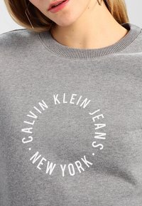 Felpa grigia realizzata in tessuto morbido. Presenta un logo circolare bianco con la scritta "CALVIN KLEIN JEANS NEW YORK" sulla parte anteriore.