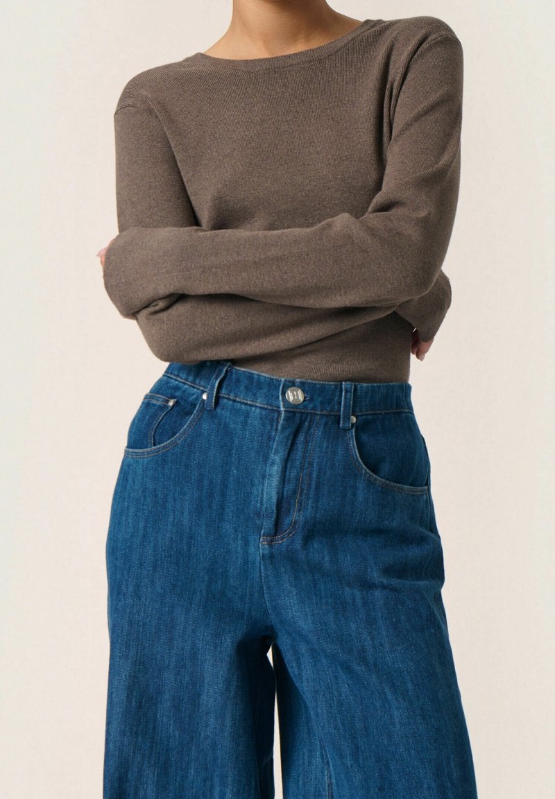 Brauner Strickpullover mit langen Ärmeln, kombiniert mit hoch taillierten blauen Denim-Jeans im weiten Schnitt, die sichtbare Taschen und einen Knopfverschluss haben.