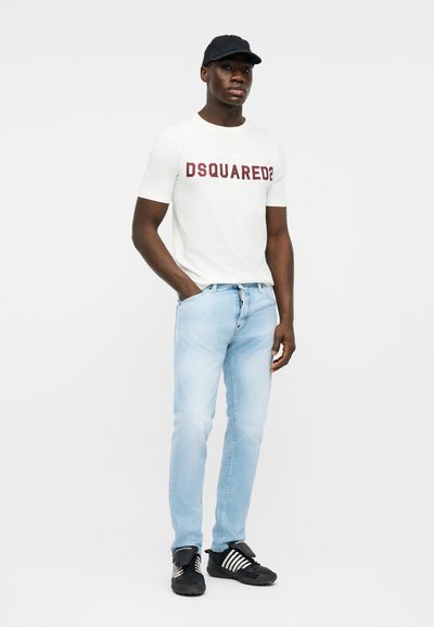Homme portant un t-shirt blanc DSQUARED2, un jean bleu clair, une casquette noire et des baskets noires à rayures blanches, debout avec une main dans la poche.