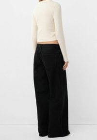 Personne portant un jean noir taille haute à jambes larges et un top court en maille côtelée crème à manches longues, debout devant un fond clair uni.