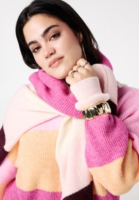 Pull en maille rayée aux teintes rose, crème et orange, avec un col montant et une texture côtelée, agrémenté d'un bracelet manchette doré.