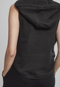 Zwarte mouwloze hoodie van zachte stof met een ontspannen pasvorm, voorzien van een capuchon en een afgeronde zoom. Zichtbare stiksels langs de naden.