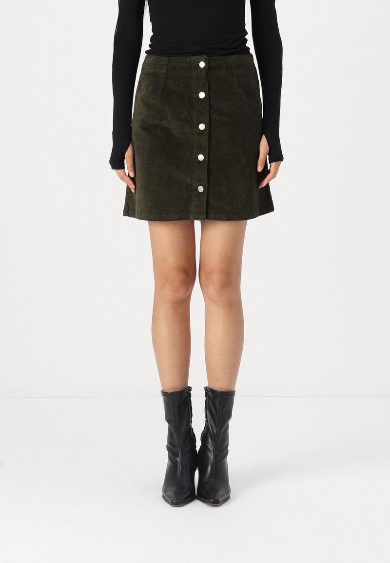 Femme portant une jupe boutonnée en velours côtelé vert foncé, un haut noir à manches longues avec des trous pour les pouces, et des bottines en cuir noir.