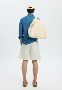 Sac fourre-tout en toile de couleur naturelle, de grande forme rectangulaire, avec une bandoulière et une poche avant. Modèle portant une chemise en denim et un short rayé.