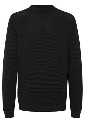 Jeune homme debout, les bras croisés, portant un pull noir, un jean bleu clair et des chaussures noires sur un fond blanc uni.
