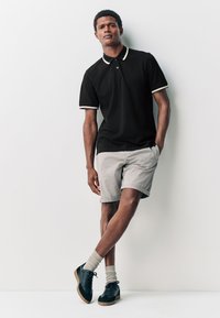 Musta poolopaita valkoisin yksityiskohdin, harmaat shortsit ja mustat kengät. Paidassa on klassinen muotoilu ja sileä pinta.