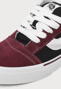Baskets Vans marron foncé et noires avec bande latérale en cuir blanc, lacets blancs, coutures visibles et semelle en caoutchouc blanc en gros plan.