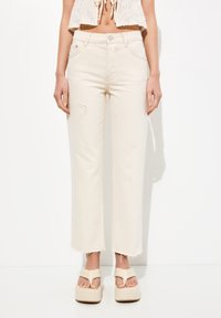 Vaqueros de mezclilla de tejido beige de cintura alta, con un corte relaxed, detalles deshilachados en la pierna izquierda y dobladillo desflecado. Combinados con sandalias de plataforma.