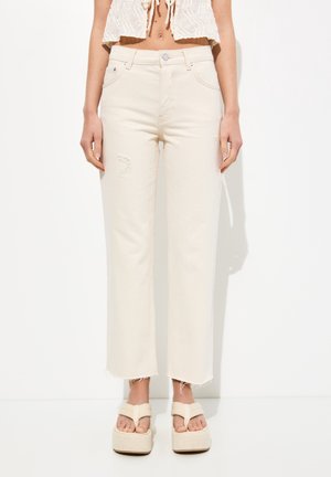 Jean beiges taille haute en denim, avec une coupe décontractée, des détails déchirés sur la jambe gauche et un ourlet effiloché. Associé à des sandales à plateforme.