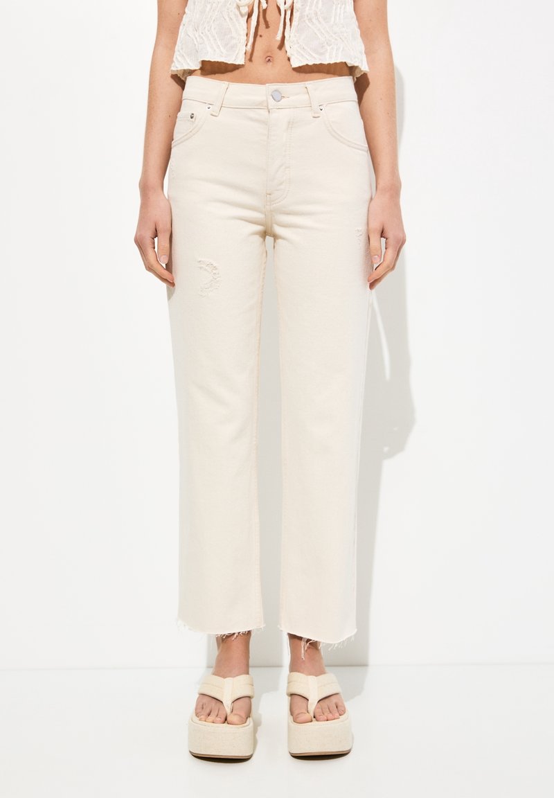 Vaqueros de mezclilla de tejido beige de cintura alta, con un corte relaxed, detalles deshilachados en la pierna izquierda y dobladillo desflecado. Combinados con sandalias de plataforma.