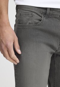 Szürke farmernadrág, slim fit szabással, standard zsebekkel, finom kopással és sima textúrával. Közeli felvétel a tenyérről a comb területén.