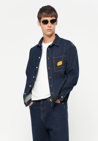 Denim jacket in dark blue with a yellow patch pocket, button closure, and gray inner lining, paired with a white t-shirt and dark denim pants.