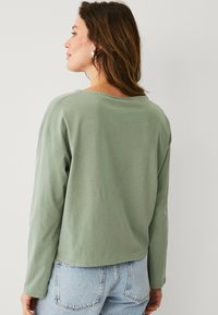 Langarmshirt aus weichem grünen Baumwollstoff, lockere Passform, Ansicht von hinten mit einem runden Halsausschnitt und leicht verkürztem Saum, kombiniert mit hellblauer Jeans.