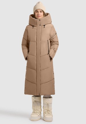 Beige langer Puffer-Mantel mit hohem Kragen, ausgestattet mit einem gesteppten Design und seitlichen Reißverschlüssen. Kombiniert mit weißen kniehohen Schneestiefeln.