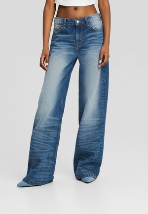 Nainen, jolla on päällään korkeavyötäröiset leveälahkeiset siniset farkut haalistunein etupaneelein ja siniset kärjest terävät kengät, näkyvissä lyhyt valkoinen toppi.