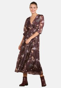 Maxikleid aus dunkellila Stoff, mit floralen Mustern, tiefem V-Ausschnitt, Puffärmeln und tailliertem Schnitt. Kombiniert mit dunkelroten Ankle-Boots.