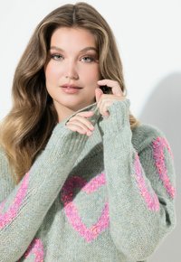 Młoda kobieta o falowanych włosach, ubrana w jasnoszary sweter z różowymi wzorami serc, poprawiająca kołnierz na białym tle.