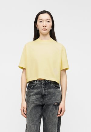 Fiatal nő hosszú fekete hajjal, laza, halványsárga rövid felsőt és magas derekú, kifakult fekete farmert visel, amelyen kis szakadás látható.