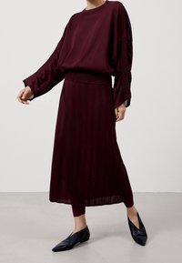 Burgundinpunainen pitkähihainen paita, jossa on teksturoidut hihat, yhdistettynä vastaavaan laskostettuun hameeseen. Asu viimeistelty tyylikkäillä mustilla kärjellään terävillä kengillä.