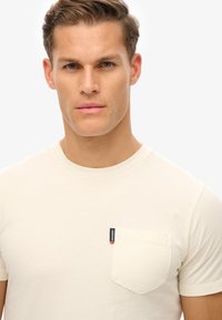 Camiseta de manga corta en color crema, hecha de una tela suave, que presenta un bolsillo en el pecho con una etiqueta de la marca en rojo y azul en el lado izquierdo.