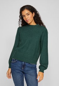 Groen gebreid trui met een ronde hals, poflange mouwen en een geribde zoom. Gekleed met blauwe jeans. Zachte textuur en een relaxte pasvorm.