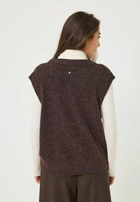Gilet tricoté brun foncé à manches courtes, avec un ourlet côtelé et un petit détail décoratif dans le dos. Porté par-dessus un col roulé léger.