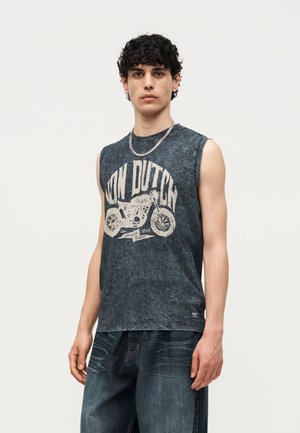 Jeune homme aux cheveux noirs bouclés portant un débardeur gris foncé avec un motif de moto et un jean ample foncé, debout devant un fond uni.