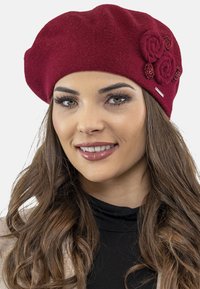 Berretto di lana rosso con superficie testurizzata, caratterizzato da tre rose in tessuto e accenti decorativi su un lato.