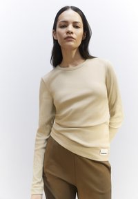 Beige, langärmliges, geripptes Oberteil mit Rundhalsausschnitt und einer subtilen Textur, kombiniert mit braunen Hosen, die ein Etikett am Bund haben.