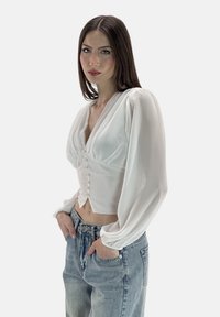Blusa bianca con maniche lunghe a sbuffo, scollo a V e sei bottoni di perla sul davanti. Abbinata a jeans in denim blu a vita alta.