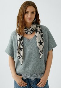 Jeune femme portant un pull en tricot gris avec une bordure en dentelle, une écharpe beige avec des motifs d'étoiles noires, et un pantalon en jean bleu, se tenant devant un fond uni.