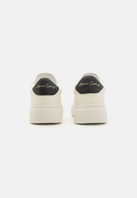 Hvit sneakers med svarte hælaksenter, laget av teksturert materiale og med et preget logo på hælen. Tykk, plattformsåle.
