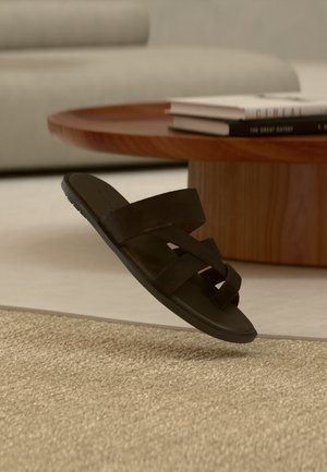 Sandale noire à bout ouvert avec des brides croisées flottant au-dessus d’un tapis beige texturé dans un salon moderne.