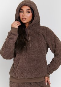 Brun fleece hoodie med en tekstureret, fluffy overflade, hætte og frontlomme. Ribstrikkede ærmer og kant i en lidt mørkere nuance.