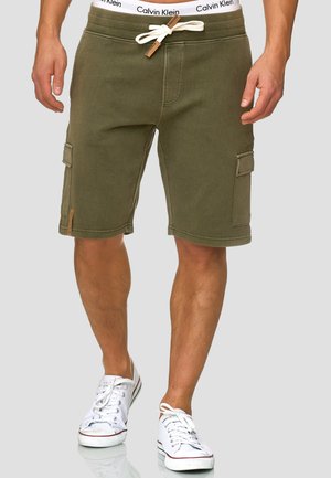 Mand iført olivengrønne cargoshorts med hvid snøre og hvide lave sneakers, stående mod en ensfarvet baggrund.