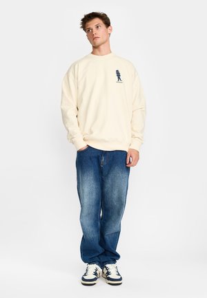 Jonge man met kort bruin haar die een crèmekleurige trui met logo, blauwe spijkerbroek en witte sneakers draagt, staand met één hand in zijn zak.