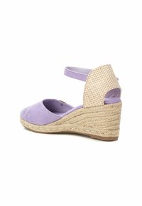 Zapato de tacón cuña de ante lila con detalles de yute trenzado en la parte posterior y correa en el tobillo; el diseño de espalda abierta permite ponérselo fácilmente.