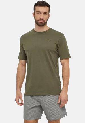 Homme debout portant un t-shirt vert olive à manches courtes et un short rayé gris sur un fond blanc uni.