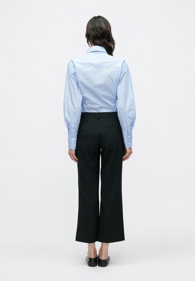 Ανοιχτό μπλε πουκάμισο με φουσκωτά μανίκια, σε συνδυασμό με μαύρο παντελόνι φαρδιάς γραμμής και μαύρα παπούτσια slip-on, εμφανιζόμενα από πίσω.