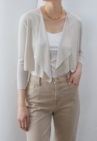 Žena oblečená v béžovém zkráceném kardiganu, bílém tílku, béžových kalhotách s vysokým pasem a masivním zlatým náhrdelníkem, drží ruku v kapse.