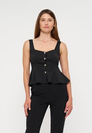Femme portant un top noir sans manches à basque avec des boutons dorés et un pantalon noir, debout devant un fond blanc uni.